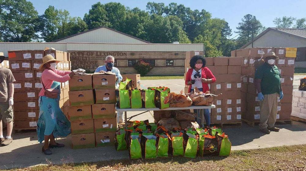 Four people work at a food pantry in front of the Lexington Multi-Purpose Complex. They are standing around a table, surrounded by many boxes and tote bags with food in them.