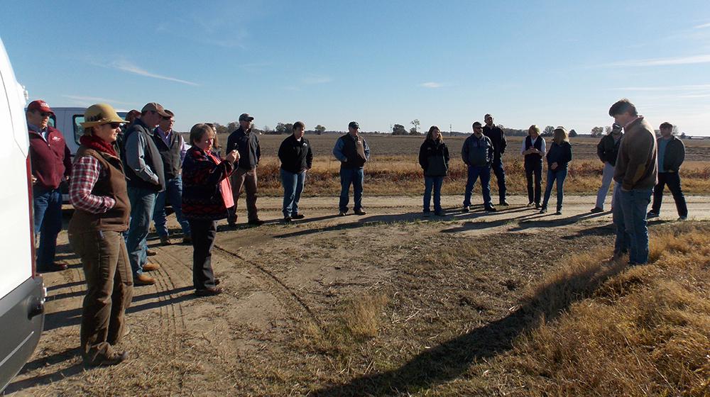 Group of people standing on dirt road on a farm listening to man giving instructions.