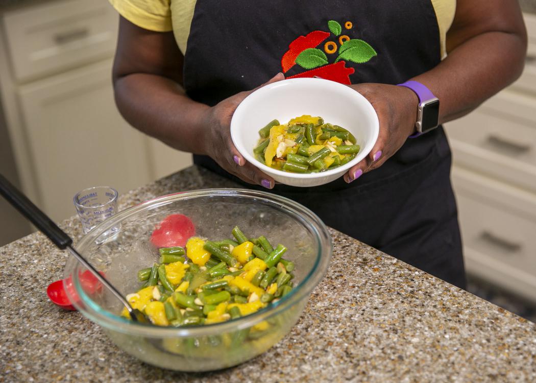 A bowl of peachy green beans sits on the kitchen counter with a woman holding a serving size in a separate bowl.