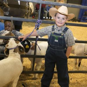 A young boy wearing overalls and a cowboy hat stands in front of a fence next to three brown and white goats.