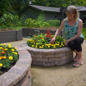 A woman wearing a blue tank top kneeling down next to a circular brick planter filled with yellow and red flowers. 