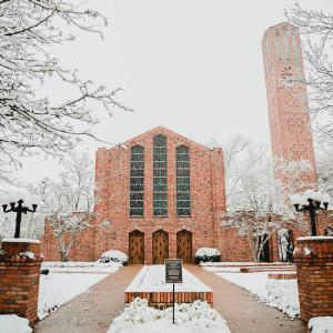 Wide view of a large brick building with three doors and three tall stained glass windows and a brick bell tower to the right.