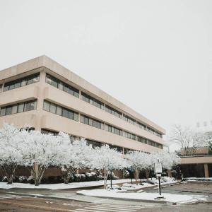 A large rectangular building surrounded by snow covered trees and an empty parking lot.