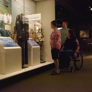 A boy, girl, and woman in a wheelchair looking at a military uniform in a museum.