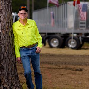 A man wearing a bright yellow button-up shirt leans against a large truck and rests one hand in his blue jean pocket.