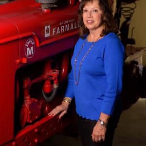 A smiling woman wearing a blue shirt stands next to and rests her arm on a red piece of machinery. 