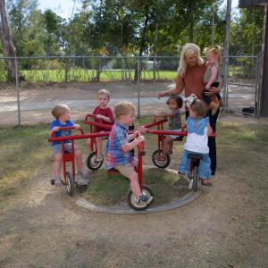 Children pedaling on a merry-go-round beside a woman holding a toddler.