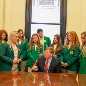 Twelve teens, standing and wearing green blazers, look at a seated man wearing a suit.