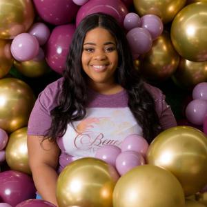 A smiling young woman wearing a shirt that reads “Beyond the Arch” stands next to several balloons and jars of party supplies.