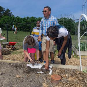 Two girls putting dirt on top of the paper towels in the raised planting bed, while a man in a plaid shirt and another student look on. 