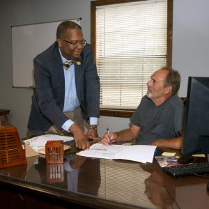 A standing Black man points to a paper on a desk that a seated white man with a pen also points to.