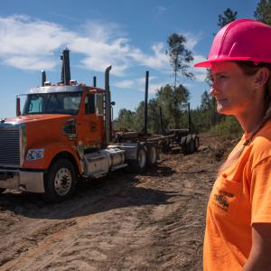 An empty, orange log hauler rolling across the cleared field as a woman wearing a hardhat looks on.