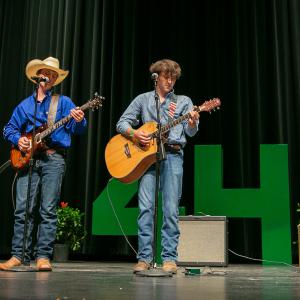 A boy wearing a cowboy hat and playing guitar sings while another boy plays also plays guitar.