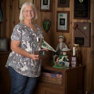 A woman, smiling, holding a 4-H flag and 4-H trophy and standing in front of several 4-H plaques affixed to the wall.