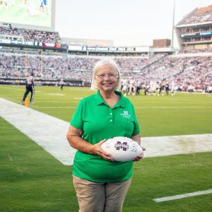 A woman wearing a green polo, standing on a football field, and holding a white football.