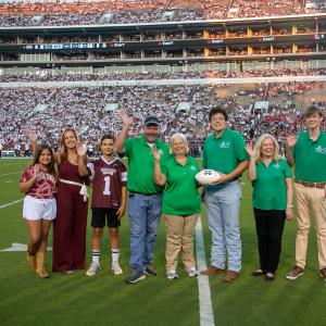 A group of people waving on a football field; the woman and man in the center holding a white football.