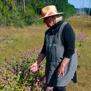 A woman with a straw hat touches purple flowers that grow waist-high.