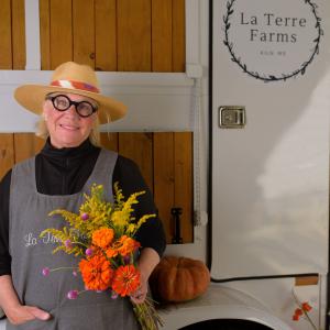 A woman with a straw hat and round-framed glasses holding a bunch of flowers and smiling.