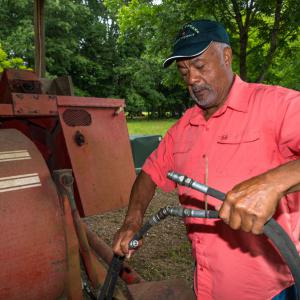A Black man holding and looking at hydraulic hoses beside an old piece of pecan equipment.