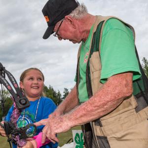 A man wearing a vest and green shirt, talking to a little girl smiling and holding an archery bow.