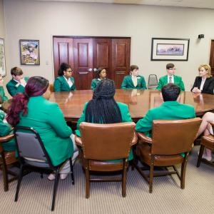 Fourteen teens in green jackets sit around a round table, and they focus on a female speaker, also sitting at the table, dressed in black jacket and white polo.