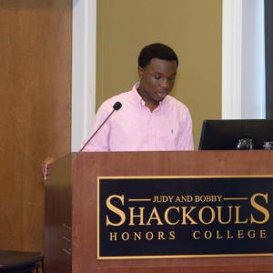A teen standing at a podium, wearing a pink shirt, speaking.