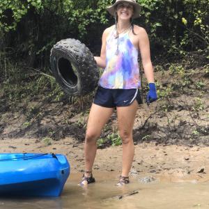 A smiling young woman wearing a colorful top stands in shallow water holding a rubber tire. 
