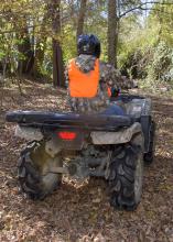 A rider wears camouflage, an orange vest and a helmet in the woods with a rifle case on the back of the ATV.
