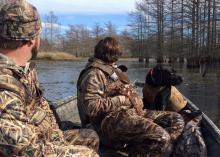 Two young men wearing camouflage sit in a small boat with a black dog, all looking out on the water on a sunny day.  