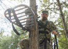 Hunter wearing camouflage secures a portable platform to the side of a tree.