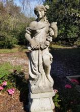Stone statue of a female with a garland of flowers stands in a flower bed.