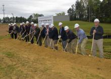 A group of more than a dozen people in hard hats break ground with shovels.
