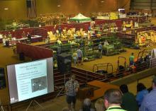 A view across an expo floor shows many individual booths and a speaker with projection screen in the foreground.