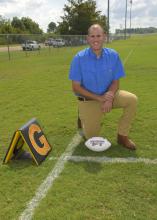 A man in a blue shirt kneels on a chalk-lined football field beside a goal line marker. A white Mississippi State football rests on the ground beside his knee.