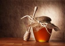 Wooden honey dipper lying against a small jar of honey that is covered with a decorative piece of fabric tied with a string.