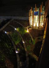Light shines at night from a cylindrical, decorative garden feature, lighting a tiered landscape bed growing reed-like vegetation near a sidewalk.
