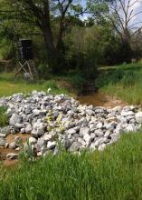 A pile of large gray rocks stretches across a ditch in a country setting.
