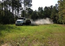 Dust billows out of a trailer on a large truck driving across a small, grassy area surrounded by tall trees.