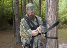Man standing in the woods inspects nylon straps on a tree stand he is holding on in his hands.