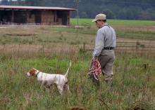 Man walks beside his bird dog on point in a pasture with a hay barn in the background.