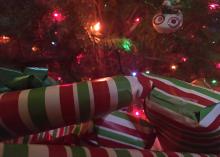 A stack of gifts wrapped in red, white and green stripes sits under a colorfully lit Christmas tree.