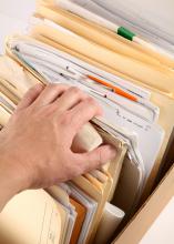 A man’s hand reaches into a box of files, pulling back the corner of one folder.