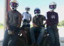 Four teenage boys wear helmets, goggles and gloves as they gather around an ATV.