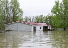 A gray, double-wide manufactured home with flood waters reaching the lower windows and surrounding area