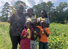 Two women and four children stand in a vegetable garden while holding yellow squash.