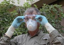 A man in a brown shirt, safety mask and rubber gloves adjusts a pair of goggles over his eyes.