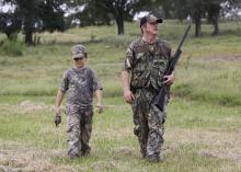 A man holding a shotgun and a boy dressed in camouflage walk in a grassy meadow.