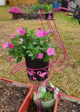 A pink flamingo made of wire holds a container with flowers.