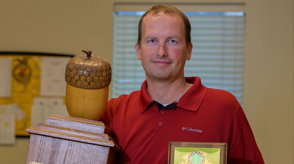 A man wearing a collared red shirt stands holding a large wooden trophy with a large wooden acorn on top in one hand and a plaque in the other.