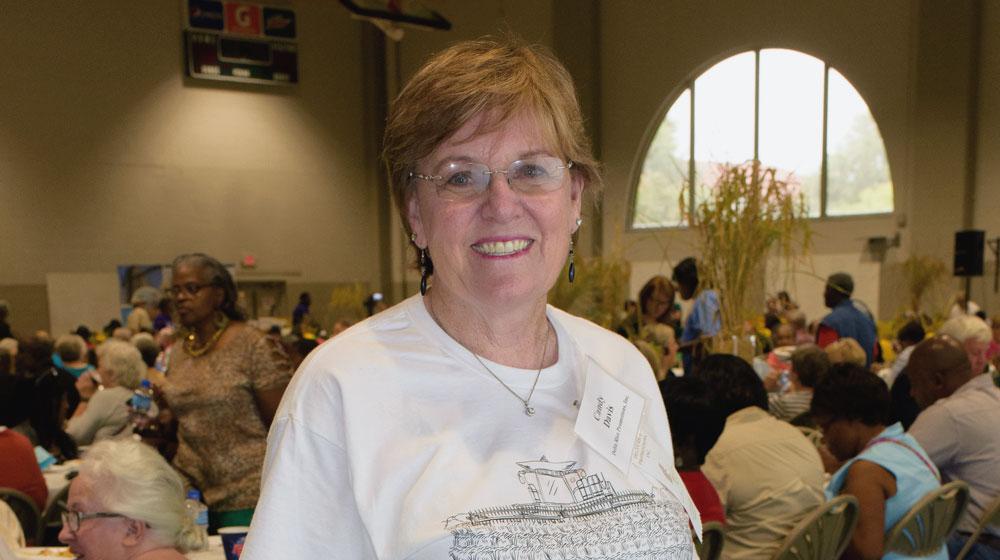 A woman stands smiling in front of a room full of people working at long tables.